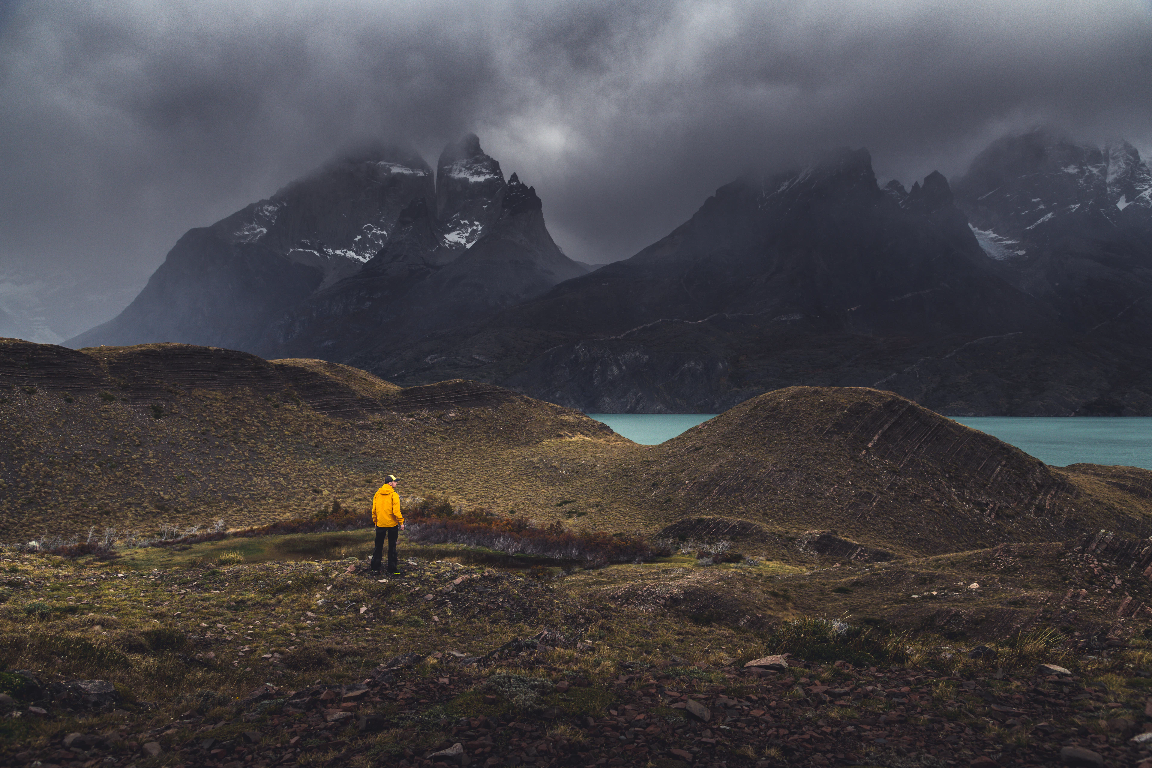 A person in a yellow jacket stands on a grassy rise below dark Patagonian peaks and a lake