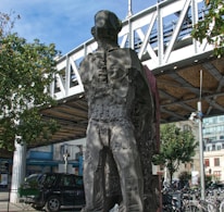 A large, abstract stone sculpture of a human figure stands beneath a metal bridge. The texture of the sculpture appears rough and eroded. Surrounding the sculpture are urban elements including a parked black car, a bicycle rack with several bikes, and nearby buildings. Trees with green foliage are visible, framing the scene and adding a natural contrast to the urban setting.