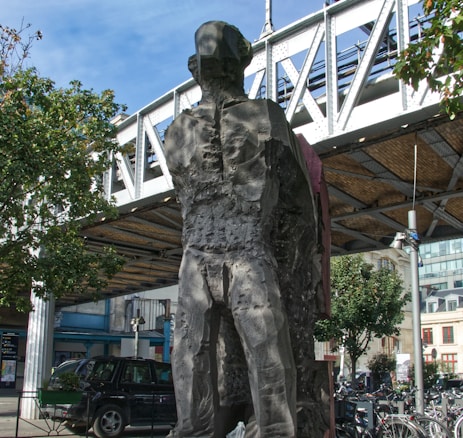 A large, abstract stone sculpture of a human figure stands beneath a metal bridge. The texture of the sculpture appears rough and eroded. Surrounding the sculpture are urban elements including a parked black car, a bicycle rack with several bikes, and nearby buildings. Trees with green foliage are visible, framing the scene and adding a natural contrast to the urban setting.