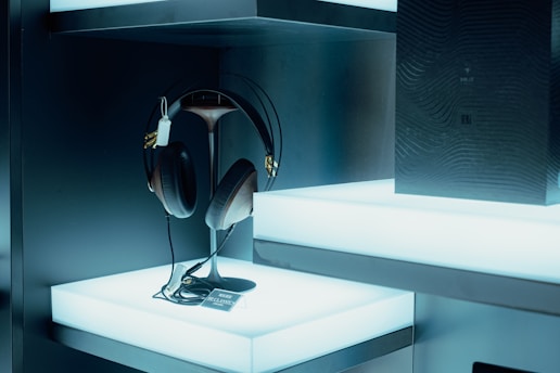 A pair of large, black and brown headphones are displayed on a sleek, modern stand. They are placed on a bright, illuminated shelf which enhances their features. Beside them, a dark, textured box with subtle design elements adds elegance to the setup. The ambient lighting is cool-toned, creating a futuristic and technologically advanced atmosphere.