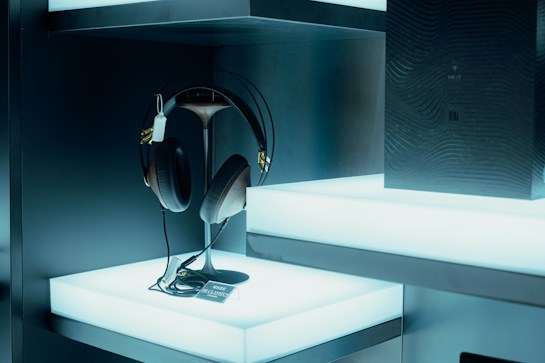 A pair of large, black and brown headphones are displayed on a sleek, modern stand. They are placed on a bright, illuminated shelf which enhances their features. Beside them, a dark, textured box with subtle design elements adds elegance to the setup. The ambient lighting is cool-toned, creating a futuristic and technologically advanced atmosphere.