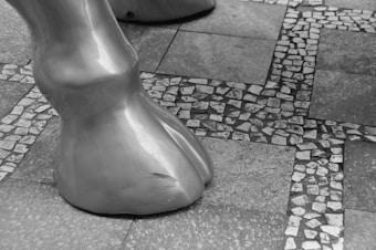 A close-up view of a large, smooth, sculpted hoof placed on a tiled mosaic floor. The floor consists of square tiles with smaller stone pieces embedded in between, creating a pattern. The image is in black and white, highlighting the textures and contrasts between the hoof and the floor.