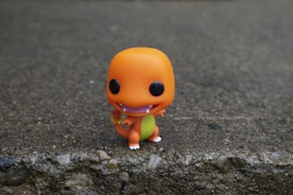 A small toy figure resembling a cartoon-like creature stands on a textured gray surface. The figure is bright orange, with an exaggerated head, large black eyes, a wide smile, and a yellowish belly. The toy is simplistic in design and has a playful appearance.
