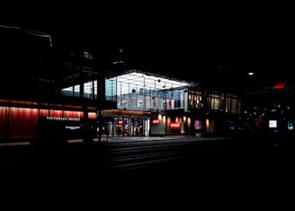 A nighttime urban scene featuring the exterior of a commercial shopping area. Storefronts of prominent brands, including Victoria's Secret and Levi's, are illuminated by bright lights. The surrounding area is dark, creating a contrast with the well-lit signs and displays. Architectural elements such as large glass windows and an overhanging structure are visible.