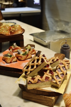 A variety of baked goods are displayed on a countertop. In the foreground, lattice-topped pastry slices filled with jam sit on a wooden tray with yellow parchment paper. Behind them, there are pastries topped with fresh fruit, including kiwi slices. The background shows a metal tray and some undefined kitchen items.
