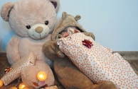 A large teddy bear and a baby dressed in a fluffy bear costume are sitting together. The baby is wrapped in a gift with polka dot wrapping paper adorned with bows, and there are warm glowing lights around.