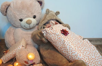 A large teddy bear and a baby dressed in a fluffy bear costume are sitting together. The baby is wrapped in a gift with polka dot wrapping paper adorned with bows, and there are warm glowing lights around.