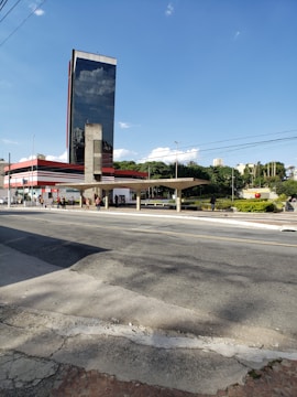 A modern office building with a reflective glass facade and red accents stands prominently against a clear blue sky. A bus stop structure with a flat roof is located in front of the building, and several people are walking along the street. The foreground features an empty road and a sidewalk, with patches of greenery in the background.