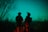 Two soldiers stand in silhouette against a night sky, with one looking through night-vision equipment. Bare trees and brush surround them in a wooded area.