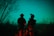 Two soldiers stand in silhouette against a night sky, with one looking through night-vision equipment. Bare trees and brush surround them in a wooded area.