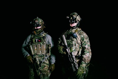 Two people wearing camouflaged military gear stand in a dark setting, each holding firearms. They are equipped with tactical vests and night vision goggles, suggesting a readiness for a nighttime operation. Their expressions and stances convey a sense of alertness and focus.