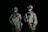 Two people wearing camouflaged military gear stand in a dark setting, each holding firearms. They are equipped with tactical vests and night vision goggles, suggesting a readiness for a nighttime operation. Their expressions and stances convey a sense of alertness and focus.
