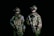 Two people wearing camouflaged military gear stand in a dark setting, each holding firearms. They are equipped with tactical vests and night vision goggles, suggesting a readiness for a nighttime operation. Their expressions and stances convey a sense of alertness and focus.