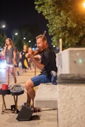 A man sitting on a bench playing a violin in an outdoor urban setting at night. The scene is lively with several people walking past, some conversing on phones. The musician is wearing a black shirt and blue shorts. Street lights create a warm, inviting ambiance.
