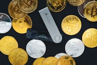 A collection of physical cryptocurrency coins scattered around a Ledger hardware wallet. The coins feature Bitcoin and Ethereum symbols, and the wallet displays a prompt to choose a PIN. The contrast between the shiny gold and silver coins and the black background creates a striking visual effect.