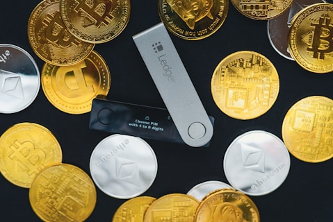 A collection of physical cryptocurrency coins scattered around a Ledger hardware wallet. The coins feature Bitcoin and Ethereum symbols, and the wallet displays a prompt to choose a PIN. The contrast between the shiny gold and silver coins and the black background creates a striking visual effect.