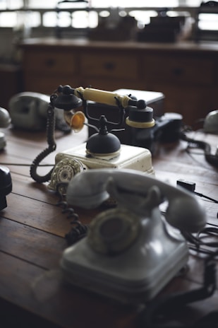 A collection of vintage rotary telephones is arranged on a wooden table. The telephones vary in color and style, with cords and dials prominently featured. The atmosphere is nostalgic, reminiscent of an earlier era of communication.