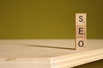 Three wooden blocks, each with a letter and a point value from a word game, are stacked vertically to spell the word 'SEO'. The blocks are placed on a wooden surface with a greenish-yellow background.