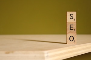 Three wooden blocks, each with a letter and a point value from a word game, are stacked vertically to spell the word 'SEO'. The blocks are placed on a wooden surface with a greenish-yellow background.