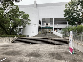 A large concrete building with a modern design and a series of steps leading up to the entrance. It is surrounded by trees and greenery. There are several signs and banners on display, and the facade exhibits clear, straight lines with minimalistic architecture.