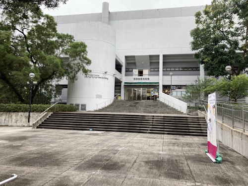 A large concrete building with a modern design and a series of steps leading up to the entrance. It is surrounded by trees and greenery. There are several signs and banners on display, and the facade exhibits clear, straight lines with minimalistic architecture.