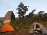 Two tents are set up on grassy terrain near an ancient stone structure resembling a temple with a Shikhara architectural style. Tall trees surround the area, with hills visible in the background. The scene depicts an outdoor camping setting in a natural and historical environment.