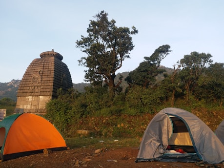 Two tents are set up on grassy terrain near an ancient stone structure resembling a temple with a Shikhara architectural style. Tall trees surround the area, with hills visible in the background. The scene depicts an outdoor camping setting in a natural and historical environment.