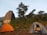 Two tents are set up on grassy terrain near an ancient stone structure resembling a temple with a Shikhara architectural style. Tall trees surround the area, with hills visible in the background. The scene depicts an outdoor camping setting in a natural and historical environment.
