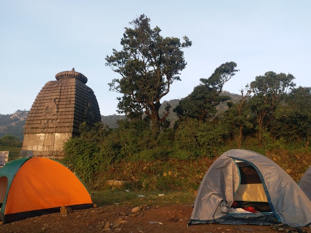 Two tents are set up on grassy terrain near an ancient stone structure resembling a temple with a Shikhara architectural style. Tall trees surround the area, with hills visible in the background. The scene depicts an outdoor camping setting in a natural and historical environment.