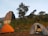 Two tents are set up on grassy terrain near an ancient stone structure resembling a temple with a Shikhara architectural style. Tall trees surround the area, with hills visible in the background. The scene depicts an outdoor camping setting in a natural and historical environment.