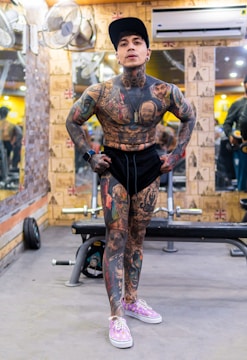 A heavily tattooed individual stands confidently in a gym setting. The person is wearing a black cap, black shorts, and pink sneakers with white laces. The background features gym equipment, including a bench press, and mirrors reflecting parts of the gym interior. The walls are decorated with wallpaper featuring various designs.