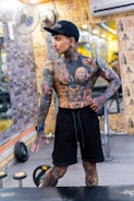 A heavily tattooed person is posing in a gym setting. The individual is dressed in black shorts and a dark cap, with an athletic build and a variety of colorful tattoos covering their body. Behind them, gym equipment such as weights and dumbbells are visible, along with mirrors lining the walls.