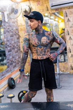 A heavily tattooed person is posing in a gym setting. The individual is dressed in black shorts and a dark cap, with an athletic build and a variety of colorful tattoos covering their body. Behind them, gym equipment such as weights and dumbbells are visible, along with mirrors lining the walls.