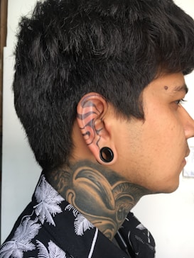 A profile view of a person with short dark hair, displaying intricate tattoos on the neck and ear. The tattoos include bold swirling patterns and designs. The person is wearing a shirt with a black and white palm tree pattern and has a large ear gauge.