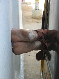 A pair of bare feet protruding from a parked vehicle, closely positioned next to a light-colored wall in an outdoor setting. The feet have a rough texture with visible cracks. The ground is sandy with some debris, and a concrete post can be seen in the background.