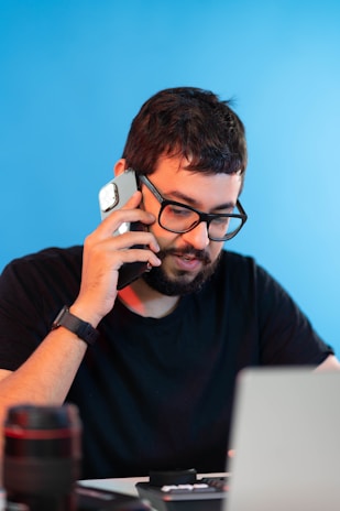 A man with glasses and a beard is sitting at a desk, holding a smartphone to his ear. He is wearing a black shirt and appears to be engaged in conversation. There is a laptop in front of him and a camera lens on the desk. The background is a solid blue color.