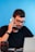 A man with glasses and a beard is sitting at a desk, holding a smartphone to his ear. He is wearing a black shirt and appears to be engaged in conversation. There is a laptop in front of him and a camera lens on the desk. The background is a solid blue color.