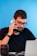 A man with glasses and a beard is sitting at a desk, holding a smartphone to his ear. He is wearing a black shirt and appears to be engaged in conversation. There is a laptop in front of him and a camera lens on the desk. The background is a solid blue color.
