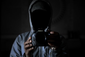A person wearing a hoodie is holding a camera, obscuring their face in shadows. The dim lighting creates a mysterious and somewhat ominous atmosphere.