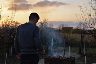 A portable BBQ grill glowing warmly with food cooking, set in a backyard at sunset.