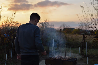 A portable BBQ grill glowing warmly with food cooking, set in a backyard at sunset.