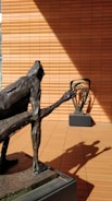 A bronze sculpture of a humanoid figure with an elongated arm is positioned in the foreground on a pedestal. The shadow of this sculpture is cast dramatically on the terracotta-colored tiled wall in the background. Another abstract sculpture is visible on a similar pedestal in the background, adding depth to the scene.