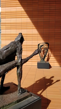A bronze sculpture of a humanoid figure with an elongated arm is positioned in the foreground on a pedestal. The shadow of this sculpture is cast dramatically on the terracotta-colored tiled wall in the background. Another abstract sculpture is visible on a similar pedestal in the background, adding depth to the scene.
