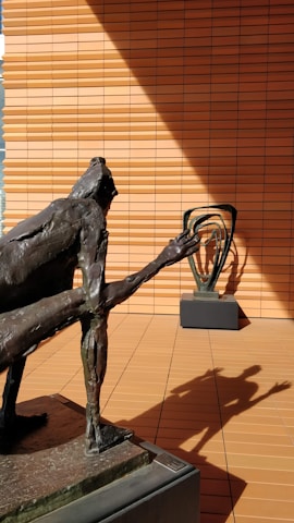 A bronze sculpture of a humanoid figure with an elongated arm is positioned in the foreground on a pedestal. The shadow of this sculpture is cast dramatically on the terracotta-colored tiled wall in the background. Another abstract sculpture is visible on a similar pedestal in the background, adding depth to the scene.