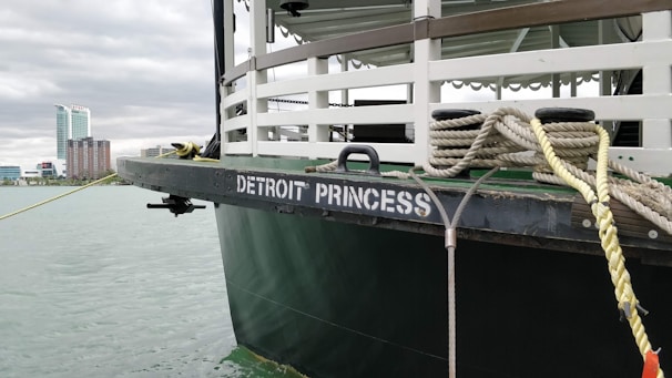 A riverboat named 'Detroit Princess' is docked near an urban waterfront. The view includes the boat's upper deck with railings and mooring ropes. In the distance, there are modern high-rise buildings and a cloudy sky.