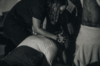 A person is huddled over another individual, possibly offering comfort or assistance. The lighting is subdued, creating a dramatic atmosphere. The image is in grayscale, enhancing the emotional intensity.