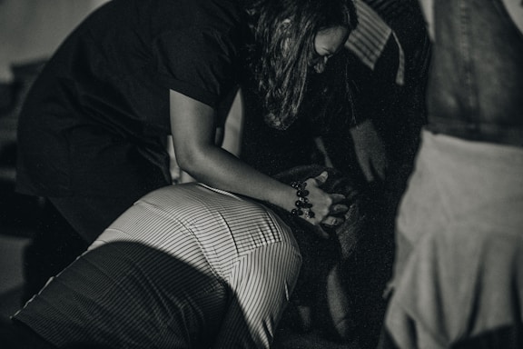 A person is huddled over another individual, possibly offering comfort or assistance. The lighting is subdued, creating a dramatic atmosphere. The image is in grayscale, enhancing the emotional intensity.