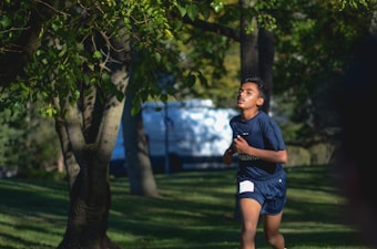 A young person is running through a lush, green park. The runner appears focused and determined, with trees and dappled sunlight creating a serene background. The person is wearing a dark sports outfit, shorts, and a t-shirt.
