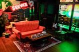 A cozy café setting features a red-orange sofa in front of a small wooden coffee table holding cups and magazines. The room is warmly lit by neon signs, with a prominent 'Service' sign above the couch. A rug with intricate patterns adds a touch of homeliness, and a guitar leans against the wall near tables in the background.
