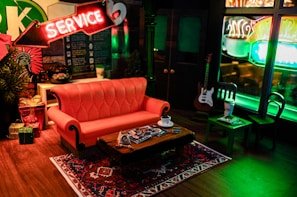 A cozy café setting features a red-orange sofa in front of a small wooden coffee table holding cups and magazines. The room is warmly lit by neon signs, with a prominent 'Service' sign above the couch. A rug with intricate patterns adds a touch of homeliness, and a guitar leans against the wall near tables in the background.